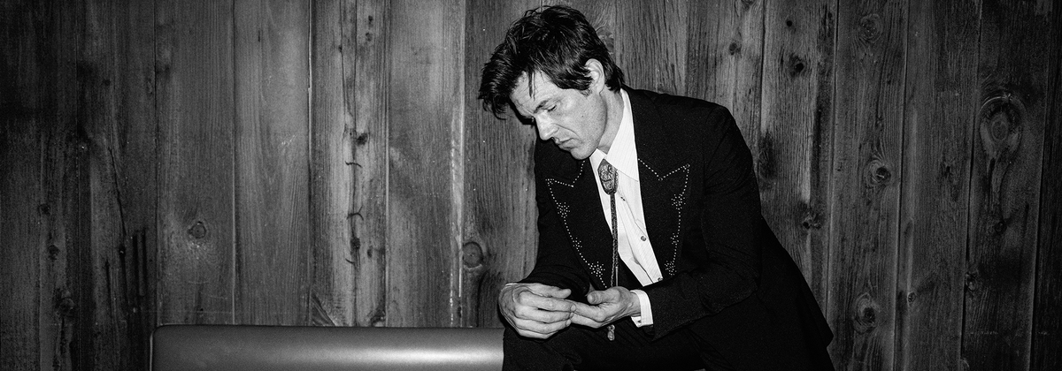 Brandon Flowers in a suit adjusting his tie against a wooden paneled wall